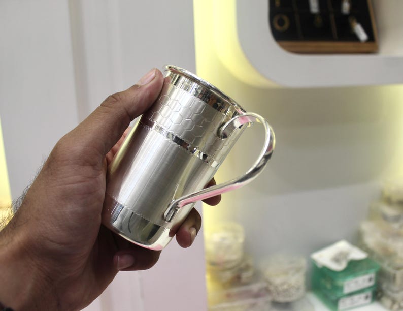 May include: A silver-colored mug with a handle. The mug has a cylindrical body with a textured pattern near the top and a smooth, brushed finish on the rest of the body. The handle is curved and attached to the side of the mug.