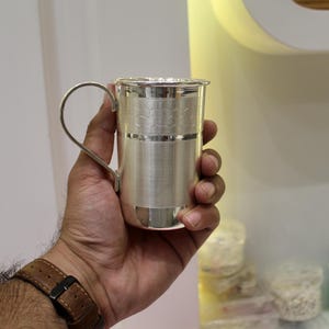May include: A silver-colored metal cup with a handle, held in a hand. The cup has a textured design around the middle and a simple handle. The cup is cylindrical and appears to be made of a shiny metal.