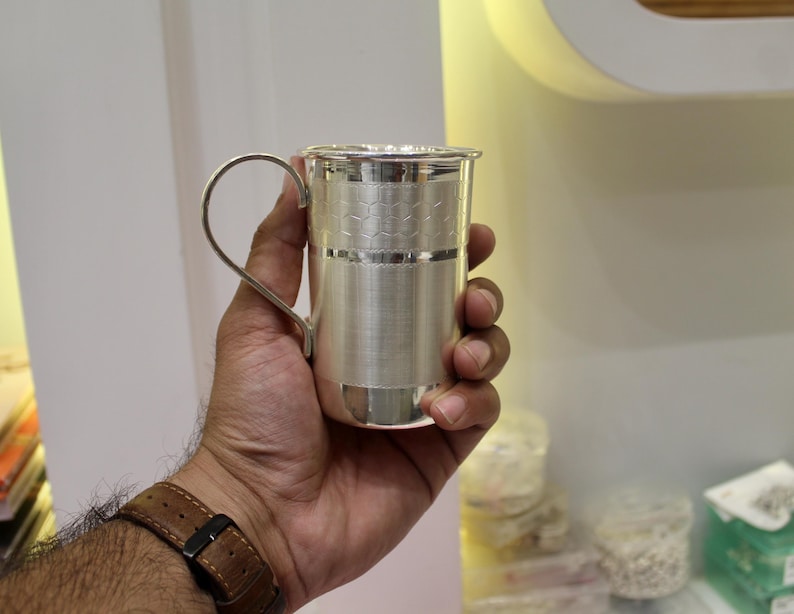 May include: A silver-colored metal cup with a handle, held in a hand. The cup has a textured design around the middle and a simple handle. The cup is cylindrical and appears to be made of a shiny metal.
