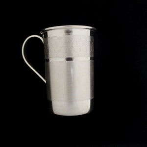 May include: A silver-colored metal mug with a curved handle. The mug has a cylindrical body with a honeycomb pattern near the top and a smooth, brushed finish on the lower portion. The mug is set against a black background.
