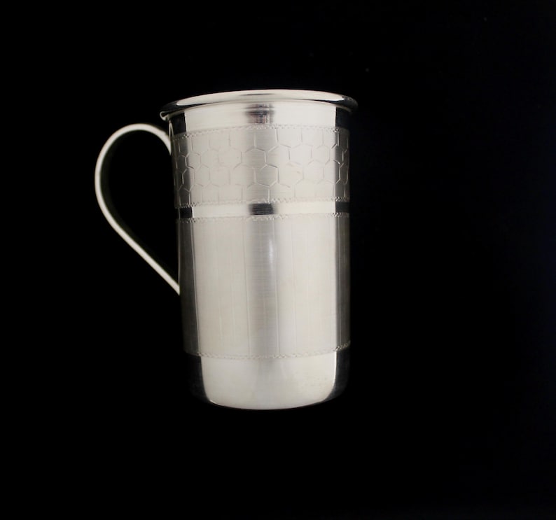 May include: A silver-colored metal mug with a curved handle. The mug has a cylindrical body with a honeycomb pattern near the top and a smooth, brushed finish on the lower portion. The mug is set against a black background.