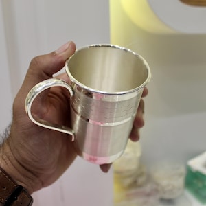 May include: A silver-colored mug with a curved handle. The mug has a textured band around the middle and a polished finish. The mug is held in a hand with a brown leather watch band visible.