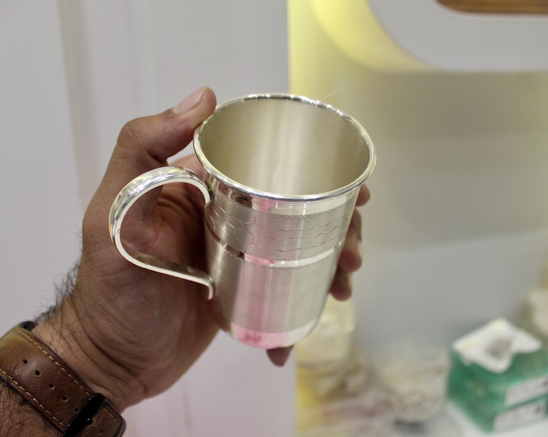 May include: A silver-colored mug with a curved handle. The mug has a textured band around the middle and a polished finish. The mug is held in a hand with a brown leather watch band visible.