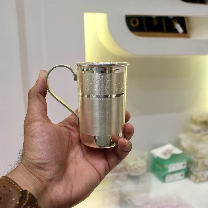 May include: A silver-colored metal cup with a handle, held in a hand. The cup has a textured design around the middle and a smooth, shiny finish. The handle is curved and attached to the side of the cup.