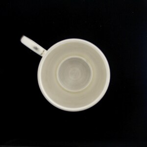 May include: Overhead view of a white ceramic mug with a handle, set against a black background. The mug's interior features a circular, recessed base. The handle is small and attached to the side of the mug. The mug appears to be empty.