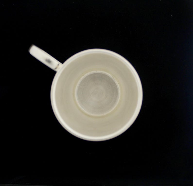 May include: Overhead view of a white ceramic mug with a handle, set against a black background. The mug's interior features a circular, recessed base. The handle is small and attached to the side of the mug. The mug appears to be empty.
