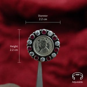 Puede incluir: Anillo plateado con un diseño central de moneda. El anillo está rodeado de piedras preciosas rojas y claras alternas. El diámetro y la altura del anillo son de 2,2 cm. El anillo es ajustable.