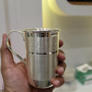 May include: A silver mug with a handle, held in a hand. The mug has a textured design with a honeycomb pattern around the middle. The mug is made of a shiny, reflective metal.