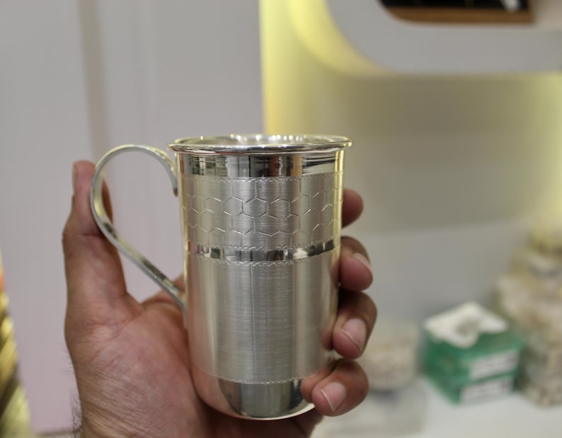May include: A silver mug with a handle, held in a hand. The mug has a textured design with a honeycomb pattern around the middle. The mug is made of a shiny, reflective metal.