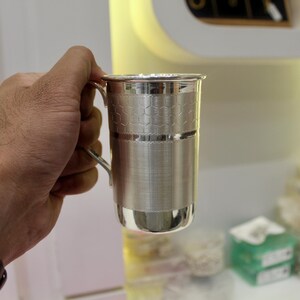 May include: A silver metal mug with a handle. The mug has a textured honeycomb pattern on the upper portion and a smooth, polished finish on the lower part. The mug is held by a person.