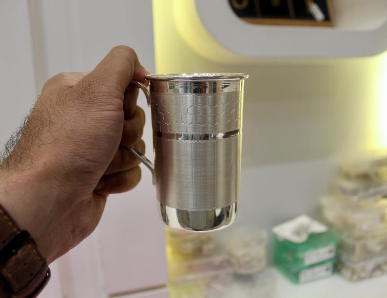 May include: A silver metal mug with a handle. The mug has a textured honeycomb pattern on the upper portion and a smooth, polished finish on the lower part. The mug is held by a person.