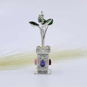 May include: A silver-colored decorative object featuring a stylized plant with green leaves. The base has a rectangular shape with ornate carvings and three colorful gemstones: pink, purple, and red. The object is set against a soft, neutral background.