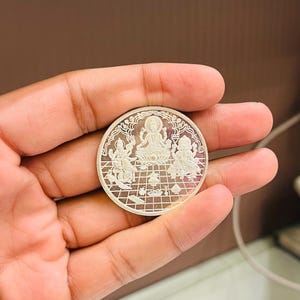 Peut inclure: Une pièce d'argent avec un motif détaillé représentant une divinité assise sur une fleur de lotus entourée de quatre autres figures. La pièce a un motif en grille sur le bord extérieur.