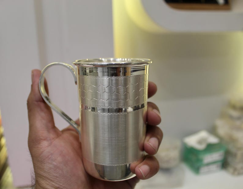 May include: A silver mug with a handle, held in a hand. The mug has a textured design with a honeycomb pattern around the middle. The mug is made of a shiny, reflective metal.