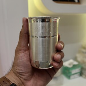 May include: A silver-colored drinking glass with a textured surface and a honeycomb pattern near the top. The glass is held in a hand, showcasing its polished finish and design. The glass is cylindrical and appears to be made of metal.