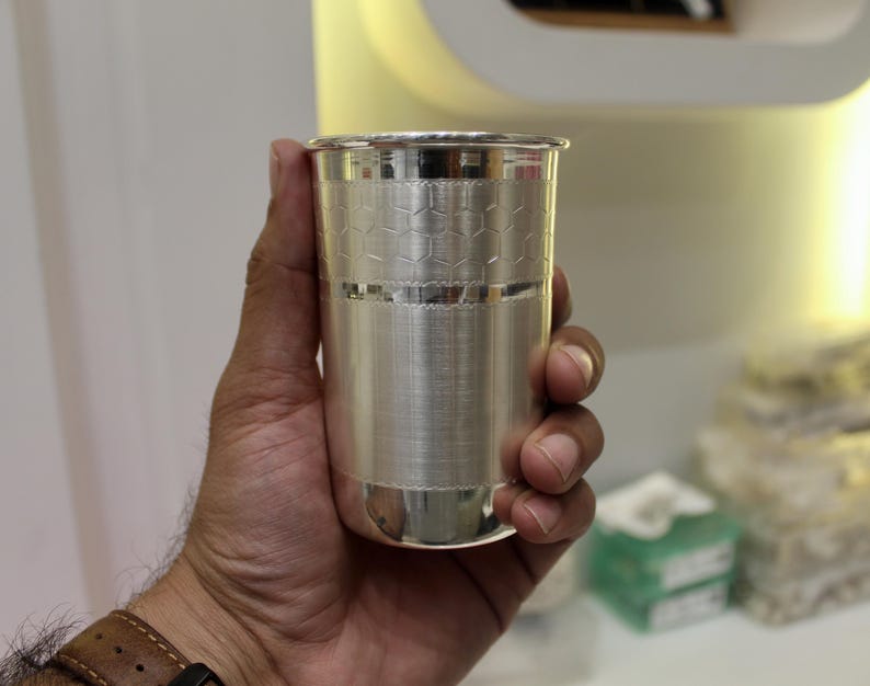 May include: A silver-colored drinking glass with a textured surface and a honeycomb pattern near the top. The glass is held in a hand, showcasing its polished finish and design. The glass is cylindrical and appears to be made of metal.