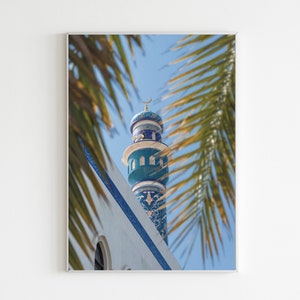 May include: A framed print showing a mosque minaret against a blue sky, with palm fronds framing the view. The minaret is decorated with turquoise and white patterns, and topped with a crescent moon. The building has a white facade with blue detailing.