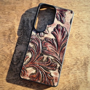 May include: A black phone case with a detailed, embossed floral pattern in shades of brown and tan. The case has a sleek, modern design with cutouts for the camera and buttons. The leather-like texture adds a touch of elegance.