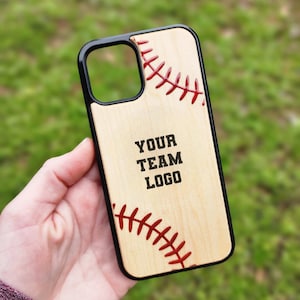 Puede incluir: Una funda de teléfono negra con un panel trasero de madera clara que presenta patrones de costuras de béisbol rojas en las esquinas superior e inferior. El centro del panel de madera muestra el texto negro "YOUR TEAM LOGO" en fuente negrita.