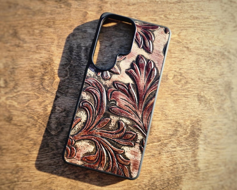 May include: A black phone case with a detailed, embossed floral pattern in shades of brown and tan. The case has a sleek, modern design with cutouts for the camera and buttons. The leather-like texture adds a touch of elegance.