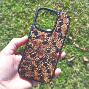 May include: A rectangular phone case with a black frame and a textured brown inlay featuring a pattern of dark, raised circular spots. The design mimics the appearance of ostrich leather with a distressed, weathered finish and a large camera cutout.