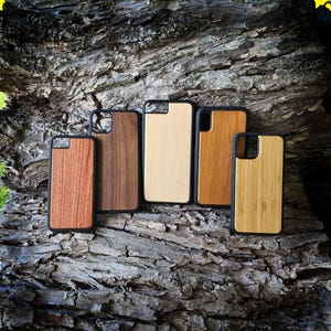 May include: Five rectangular phone cases with black protective edges and distinct wood grain back panels in varying shades of light, medium, and dark brown, arranged in a row. Each case features a cutout for a camera lens on the top corner.