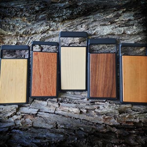 May include: Five rectangular phone cases with black frames and distinct wood grain back panels in varying shades of light, medium, and dark brown. Each case features a horizontal cutout at the top for a camera module, showcasing natural wood textures.