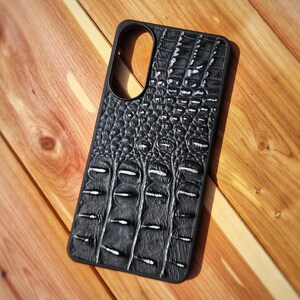 May include: A black smartphone case featuring a textured, raised pattern that mimics the appearance of crocodile skin. The case has a rectangular shape with rounded corners and a large, oval-shaped cutout at the top for a camera lens assembly.