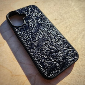 Puede incluir: Una funda de teléfono negra con un patrón floral en relieve en toda la superficie posterior. La funda incluye un recorte circular en la esquina superior izquierda para el lente de la cámara y mantiene un color oscuro constante.