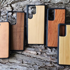 May include: Five rectangular smartphone cases with black protective frames and inlaid wood panels in varying shades of light, medium, and dark brown. Each case features a cutout for a camera lens array on the upper left or centre back.
