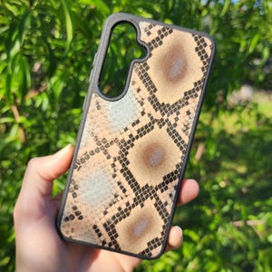 May include: A rectangular smartphone case featuring a black frame and a textured snakeskin pattern in shades of tan, brown, and light blue. The design includes a large cutout at the top for a camera lens and a black border surrounding the entire case.