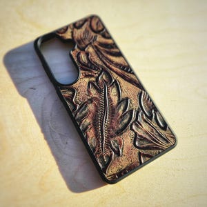 May include: A rectangular phone case with a black frame and a textured, embossed surface featuring a brown and tan leaf pattern. The design includes deep, carved-looking details of foliage across the back panel, with a cutout for a camera lens.