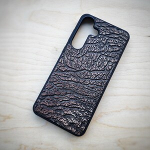 May include: A rectangular phone case with a black border and a textured, dark brown surface featuring a raised, irregular pattern resembling elephant skin. The case includes a large, rounded cutout at the top for a camera lens assembly.