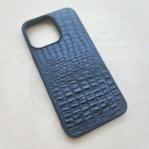 Puede incluir: Una funda para smartphone de color negro con un patrón texturizado de piel de cocodrilo en la superficie posterior. La funda incluye un gran recorte rectangular en la esquina superior izquierda para el módulo de cámara y un borde elevado.