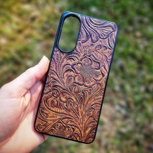 May include: A rectangular phone case featuring a brown, embossed floral pattern with intricate swirling leaves and petals. The design has a dark, antiqued finish within the recessed areas, contrasting with the lighter brown raised surface details.