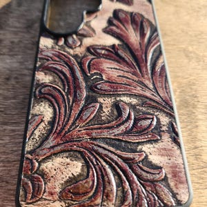May include: A phone case with a black frame and a detailed, embossed leather design. The leather features a floral pattern in shades of brown and burgundy, with a textured, aged appearance.