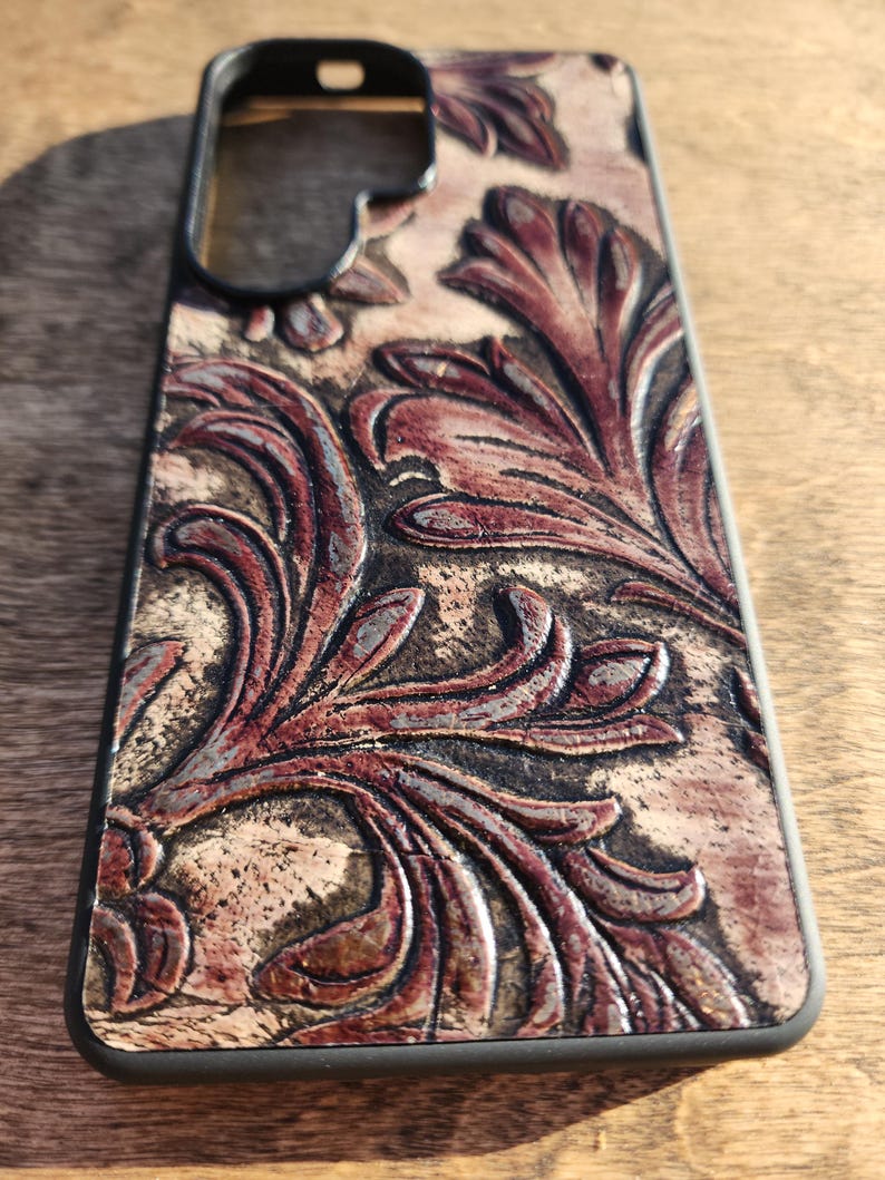 May include: A phone case with a black frame and a detailed, embossed leather design. The leather features a floral pattern in shades of brown and burgundy, with a textured, aged appearance.