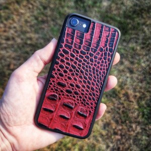 May include: A smartphone case featuring a textured, deep red and black crocodile skin pattern. The case has a black frame and a cutout for the rear camera lens and flash. The design covers the entire back surface of the mobile device.