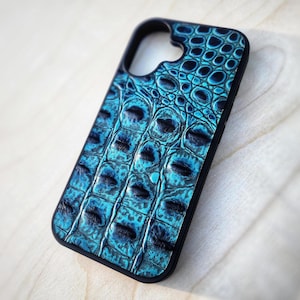 Peut inclure: Une coque de téléphone rectangulaire arborant un motif texturé en peau de crocodile bleu sarcelle et noir. L'étui présente un contour noir et une grande découpe circulaire en haut pour l'objectif, avec un relief détaillé en écailles.