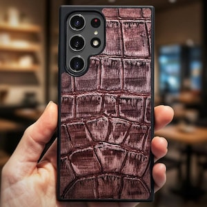 May include: A rectangular smartphone case featuring a dark brown, textured crocodile skin pattern. The case has a black frame and a cutout section for the rear camera array, which contains four circular lenses and a small flash component.