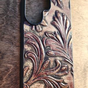 May include: A phone case with a black frame and a brown, embossed leather design. The leather features a detailed floral pattern in shades of brown and tan, creating a textured, vintage aesthetic. The case is designed to protect a smartphone.