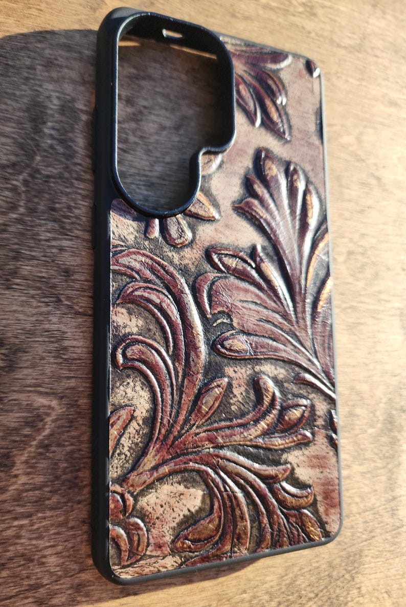May include: A phone case with a black frame and a brown, embossed leather design. The leather features a detailed floral pattern in shades of brown and tan, creating a textured, vintage aesthetic. The case is designed to protect a smartphone.