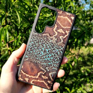 May include: A rectangular smartphone case with a black frame and a textured back panel featuring a brown and turquoise snakeskin-inspired pattern. The case includes a large cutout at the top for a camera array and follows a slim, rigid design.