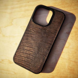 May include: A rectangular phone case featuring a black frame and a textured brown back panel with a detailed embossed pattern that mimics the appearance of reptile skin. The case includes a large cutout at the top for a camera lens array.
