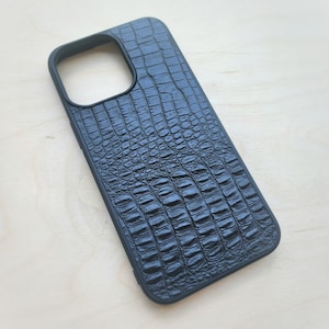 May include: A black smartphone case featuring a textured crocodile skin pattern across the back surface. The case includes a large rectangular cutout at the top left corner for a camera module and has a solid black frame surrounding the edges.