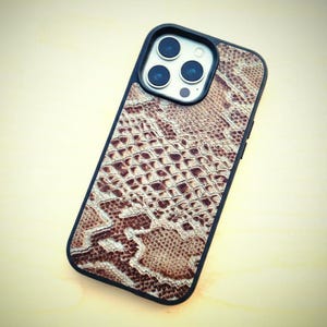 May include: A smartphone case featuring a brown and beige snakeskin pattern with a black border. The case has a cutout for a triple-lens camera system on the upper left corner, showing three circular lenses and a flash component.