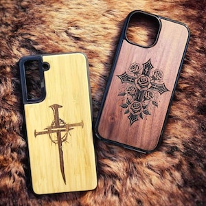 May include: Two rectangular phone cases with black protective borders. The left case is light-colored wood featuring a laser-engraved design of three nails arranged as a cross with a crown of thorns. The right case is dark wood with a cross and rose design.