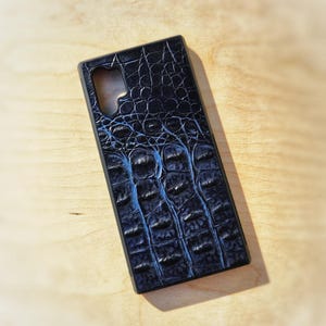 Peut inclure: Une coque de téléphone rectangulaire présentant un motif texturé en peau de crocodile bleu foncé et noir. La coque possède une découpe distincte dans le coin supérieur gauche pour l'objectif de l'appareil photo et un cadre rigide.