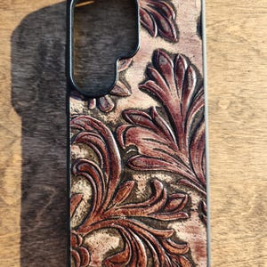 May include: A brown leather phone case with a black border. The case features a detailed, embossed floral design in shades of brown and tan. The design is centered on the back of the case.