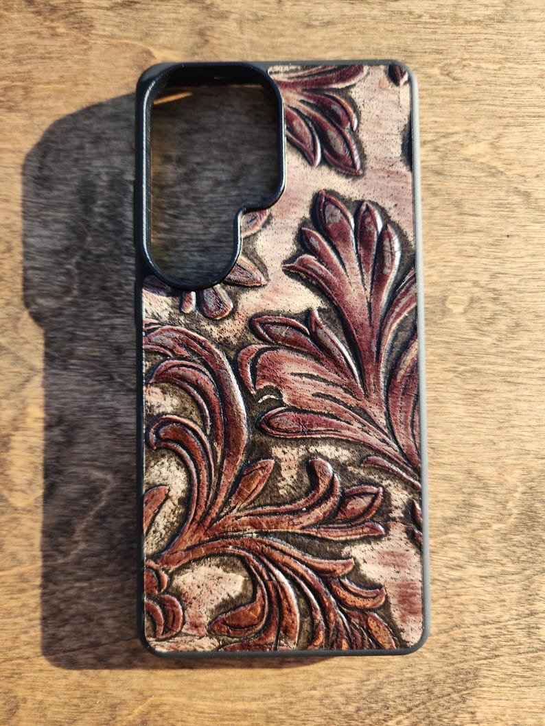 May include: A brown leather phone case with a black border. The case features a detailed, embossed floral design in shades of brown and tan. The design is centered on the back of the case.